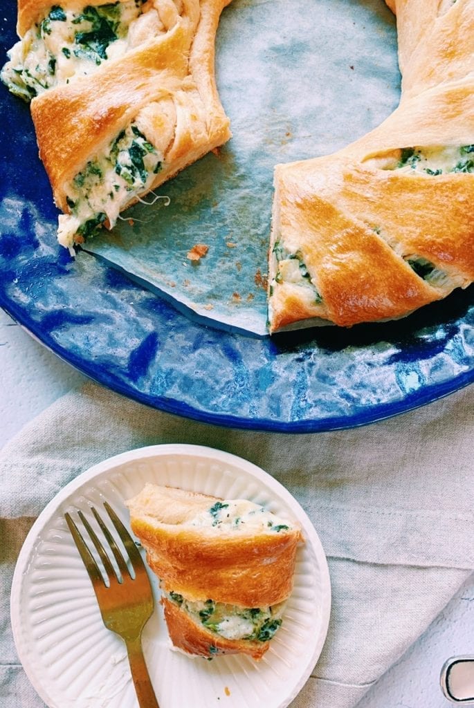 Slice of spinach and artichoke dip ring on a white plate with the rest of the ring on a blue plate next to it. 