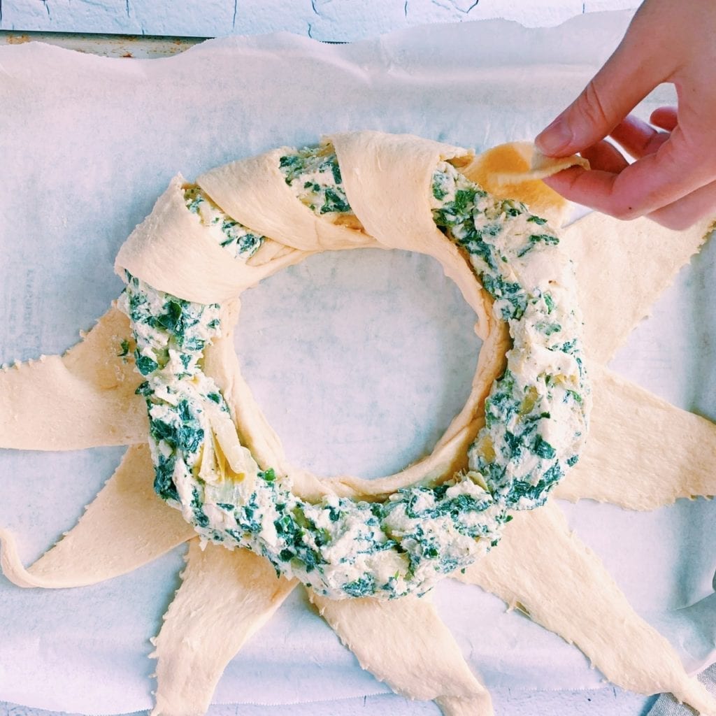 A hand pulling the tip of a crescent roll triangle over spinach and artichoke dip ring. 