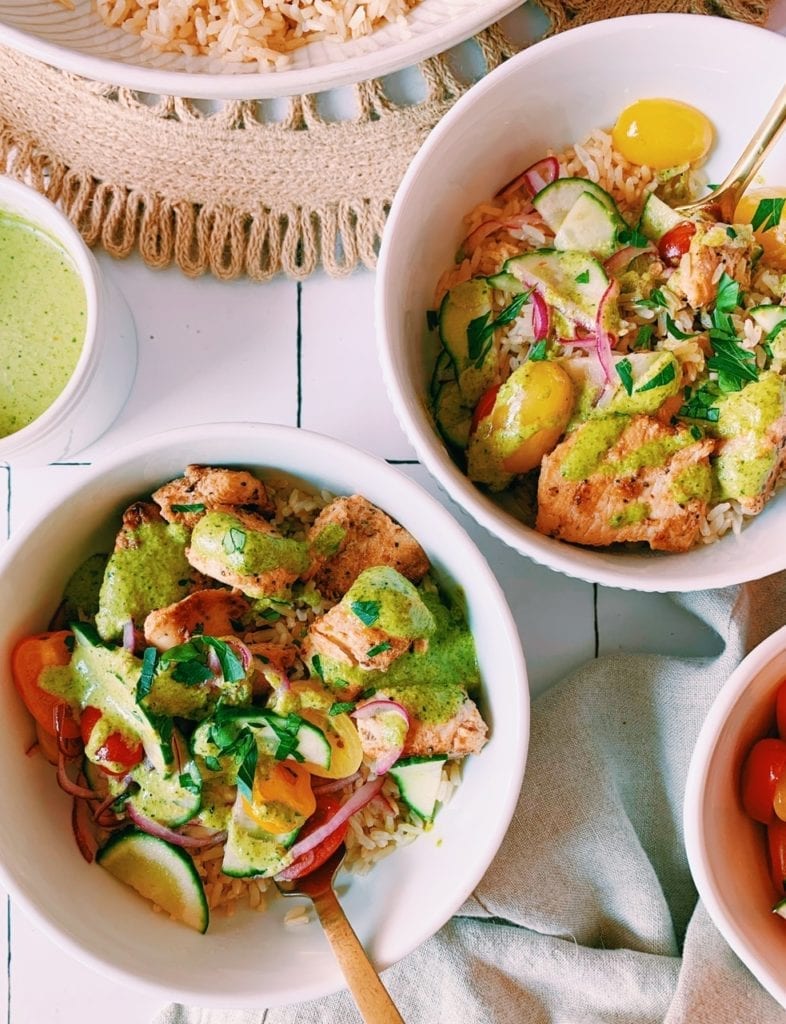 Flatlay of mediterranean rice and chicken bowls with cucumber tomato salad and spicy green sauce in white bowls. 