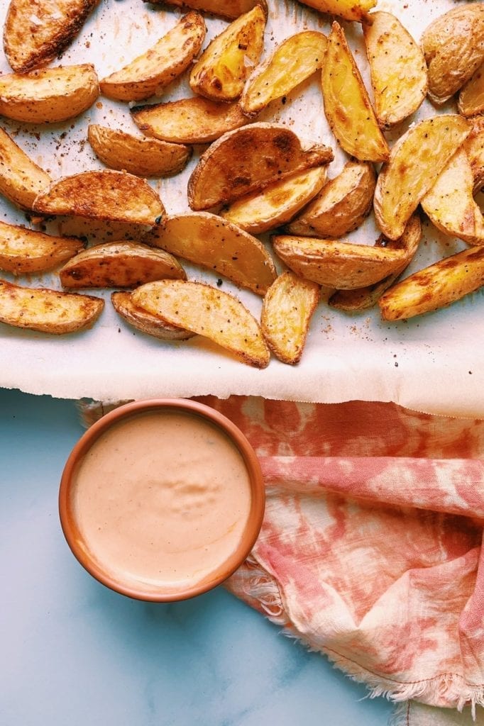 The yummiest potato wedges on a parchment-lined baking sheet with chipotle dip in a small dish next to it. 