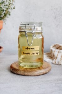 thyme simple syrup in a clear glass jar sitting on a white coaster