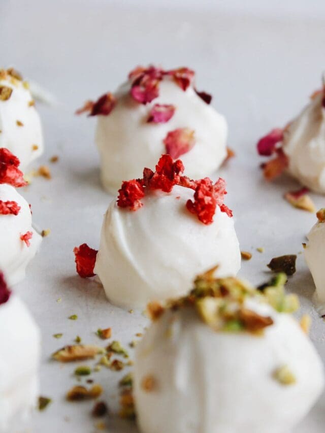 rose white chocolate truffles on a baking sheet topped with chopped pistachio, strawberries and rose petals.