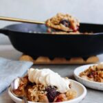 cherry apple crumble on a white plate with cast iron pan behind it