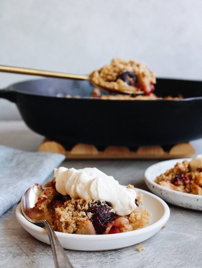 cherry apple crumble on a white plate with cast iron pan behind it