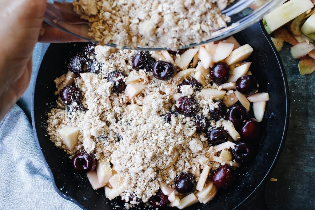 cherry apple crumble topping being poured over fruit filling in cast iron skillet